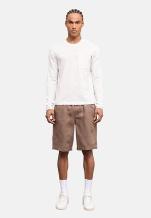 Uomo con capelli intrecciati che indossa una camicia bianca a maniche lunghe con taschino sul petto, pantaloncini marroni fino al ginocchio, calzini bianchi e scarpe da ginnastica bianche, che sta fermo.