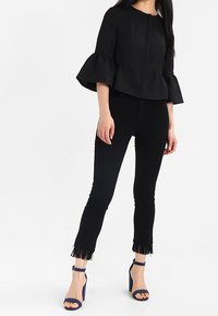 Zwarte, textuur peplumtop met uitlopende mouwen, gecombineerd met strakke zwarte jeans met franjes aan de onderkant en marineblauwe hoge hakken.