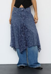 Személy rétegezett kék csipke szoknyát visel, széles szárú farmernadrág felett, fekete hegyes cipőben, egy sima fehér falnak támaszkodva.