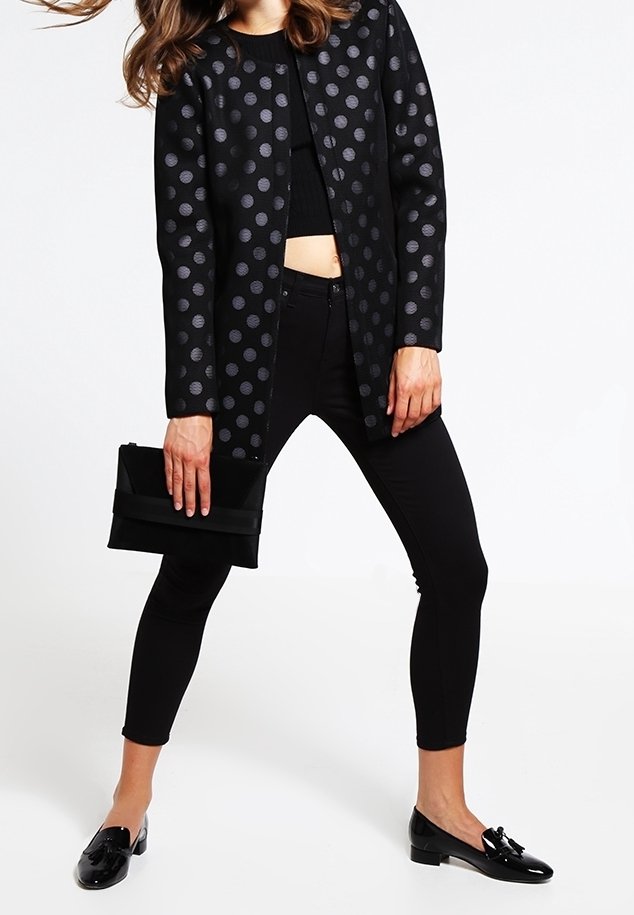 Femme portant une veste noire à pois, un top court noir, un pantalon slim noir, des mocassins noirs, tenant une pochette noire.