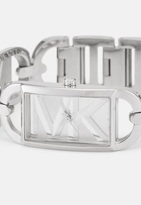 Stříbrné nerezové ocelové náramkové hodinky s obdélníkovým ciferníkem, který má texturovaný vzor, výrazné logo "MK" a design propojeného náramku.