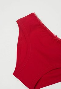 Rote Baumwollslips mit glatter Textur, versehen mit einem gewellten Saum und einem kleinen Schleifenakzent auf der linken Vorderseite.