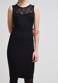 Vestito nero senza maniche con un corpetto strutturato caratterizzato da dettagli in pizzo floreale e una gonna a coste aderente, che termina appena sotto il ginocchio.