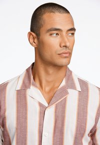 Camisa de botones de manga corta con rayas verticales en tonos de burdeos, crema y melocotón. Presenta un cuello plano y una tela ligera.