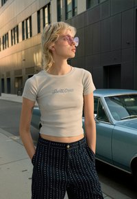 Világosszürke, rövidített fazonú póló "Santa Cruz" felirattal; sötétkék nadrággal, fehér függőleges csíkokkal és szívmintákkal. Rózsaszín napszemüveg.