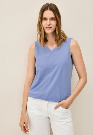 Top blu, senza maniche, con scollatura a V e texture a coste, abbinato a pantaloni bianchi; il modello posa con una mano nella tasca su uno sfondo neutro.
