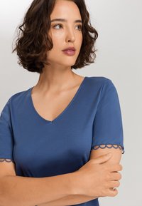 Top blu con scollo a V e maniche corte, caratterizzato da dettagli smerlati sui bordi delle maniche. Tessuto liscio e silhouette aderente.