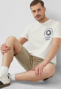 Witte katoenen t-shirt met een marineblauw grafisch ontwerp, gecombineerd met lichtkhaki shorts en witte sneakers met zwarte accenten, met enkelsokken.