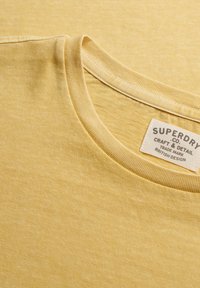 Close-up van een bleekgele T-shirt met ronde hals en een wit stoffen label met de tekst "Superdry Co. Craft & Detail Trade Mark British Design."
