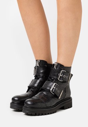 Botines negros con textura de cocodrilo, suelas gruesas y dos grandes hebillas plateadas, usados en piernas desnudas sobre un fondo blanco.