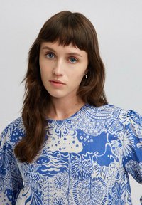 Sinine ja valge mustriga pluus ümmarguse kaelusega ja puffse varrukatega, millel on lille- ja abstraktsed kujundid tekstuuritud kangal.