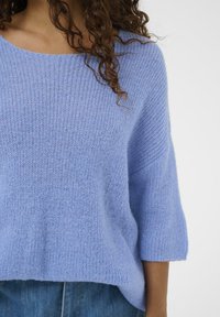 Pull en tricot bleu clair, coupe ample, encolure large et manches trois-quarts. Texture douce avec détails côtelés à l'ourlet.