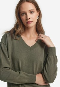 Olivgrüner V-Ausschnitt-Pullover aus weichem, strukturiertem Stoff mit lockeren Ärmeln. Auffällige gerippte Akzenten am Kragen und an den Bündchen.
