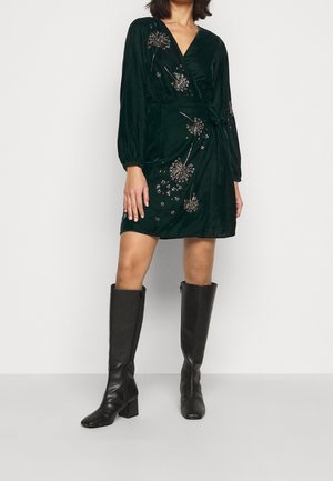 Kobieta ubrana w ciemnozieloną aksamitną sukienkę kopertową z kwiatowym haftem oraz czarne, sięgające kolan, na obcasie kozaki, stojąca na jednolitym tle.