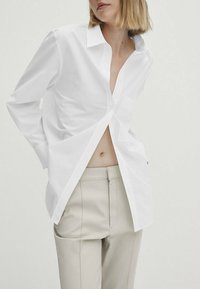 Chemise blanche à manches longues avec un col pointu, boutonnée à l'avant et un design ouvert, associée à un pantalon beige sur mesure présentant une texture lisse.