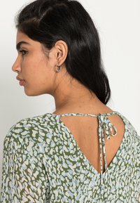 Top verde com padrão e costas em V, com fecho de laço. O design inclui detalhes em azul e branco, tecido suave e mangas curtas.