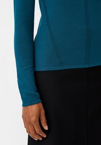 Türkises Langarmshirt aus weichem Stoff, mit strukturierten Nähten und tailliertem Design, kombiniert mit einem schwarzen Kleidungsstück.