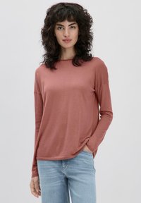 Langärmliges, mauves Oberteil mit lockerer Passform, rundem Ausschnitt und weicher Textur. Kombiniert mit hellblauen Jeans. Dezente Seitennahtdetails.