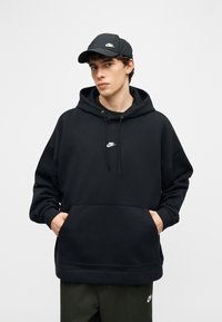 Νέος άνδρας που φοράει μαύρο φούτερ με κουκούλα Nike και μαύρο καπέλο, με τα χέρια στην τσέπη του φούτερ, στέκεται μπροστά σε απλό ανοιχτό γκρι φόντο.