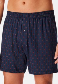 Marineblå boxershorts lavet af blødt stof med et geometrisk mønster af røde og sorte linjer. Har en elastisk talje og en enkelt knap til lukning.