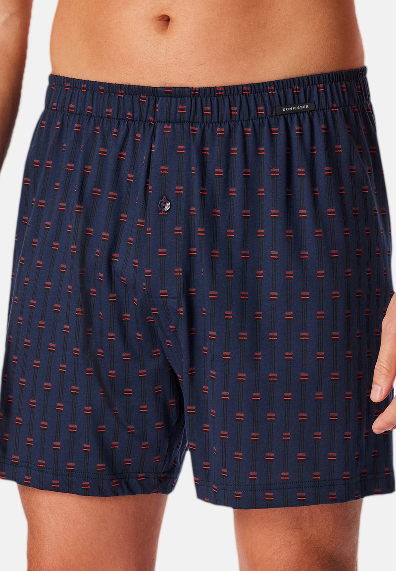 Marineblå boxershorts lavet af blødt stof med et geometrisk mønster af røde og sorte linjer. Har en elastisk talje og en enkelt knap til lukning.