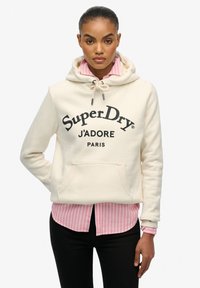 Crème kleurige hoodie met de tekst "SuperDry J'ADORE PARIS" in zwart, voorzien van een trekkoord in de capuchon en een buidelzak aan de voorkant, gelaagd over een roze gestreept shirt.