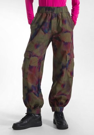 Osoba nosząca wielokolorowe spodnie cargo tie-dye z elastycznymi ściągaczami, czarne trampki i jasnoróżową bluzkę z długim rękawem, ręce w kieszeniach.