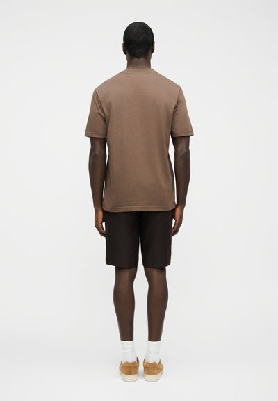 Άνδρας που φορά καφέ κοντομάνικο πουκάμισο, σκούρο σορτς, άσπρες κάλτσες και μπεζ αθλητικά παπούτσια, στέκεται πλάτη σε λευκό απλό φόντο.