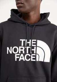 Zwarte hoodie van een katoenmix, met een groot wit logo-ontwerp "THE NORTH FACE" en een halfcirkelvormig design op de voorkant. Verstelbare capuchon.