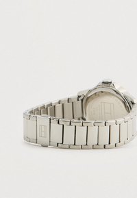 Sølvmetal kædearmbånd med spænde og et emblem, fastgjort til et rundt urkasse med en synlig krone mod en hvid baggrund.