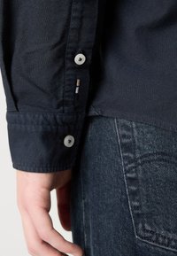 Gros plan d'une main à côté d'un poignet de chemise bleu foncé boutonné et d'un jean en denim bleu avec poche arrière visible et coutures apparentes.