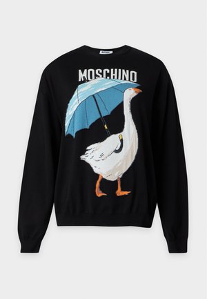 Kobieta w czarnej bluzie Moschino z białą gęsią trzymającą niebieski parasol, zestawiona z luźnymi szarymi spodniami i czarnymi sandałami.