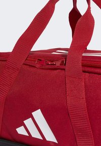 Bolsa de deportes de tela roja con cierre superior, que presenta el logotipo blanco de Adidas y asas trenzadas para llevar. Detalle de superficie texturizada visible.