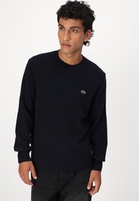 Σκούρο μπλε πουλόβερ φτιαγμένο από πλεκτό ύφασμα με στρογγυλή λαιμόκοψη, ριμπ μανσέτες και στρίφωμα, με ένα μικρό πράσινο λογότυπο κροκοδείλου στο στήθος.