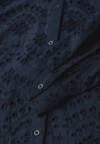 Camisa azul marino con patrones bordados intrincados y detalles recortados. Cuenta con dos botones en la parte frontal y una manga doblada de forma ordenada.