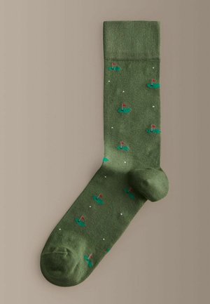 Zelená ponožka s malými motívmi golfových jamiek obsahujúcimi červené zástavky a biele golfové loptičky rozptýlené po tkanine.
