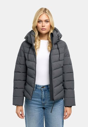 Frau mit langen blonden Haaren trägt eine graue wattierte Kapuzenjacke über einem weißen Hemd und blauen Jeans, schaut nach vorne mit neutralem Gesichtsausdruck.