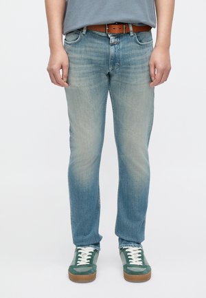 Jeans en denim bleu clair avec une finition délavée, coupe droite, style cinq poches, et une ceinture en cuir marron. Portés avec des baskets vertes.
