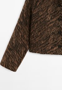 Cardigan marron texturé avec une finition duveteuse, présentant un design court et des boutons noirs. Le tissu affiche un motif chevron subtil.