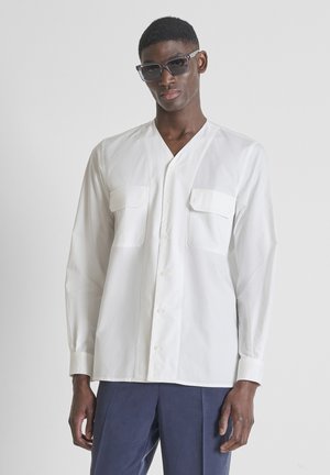 Mannelijk model met een zonnebril, een wit overhemd met lange mouwen en borstzakken, en donkerblauwe broek, staand tegen een effen achtergrond.