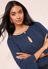 Marineblauw top met lange mouwen en een ronde halslijn met een keyhole-detail, gemaakt van gestructureerde stof en voorzien van een decoratieve knoop bij de kraag.
