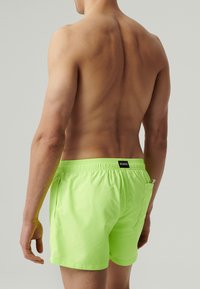 Neonowe zielone szorty kąpielowe z elastycznym pasem, bocznymi kieszeniami oraz tylną kieszenią z małą metką z logo. Teksturalny, lekki materiał.