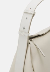 Borsa a mano strutturata di colore beige con una larga tracolla, realizzata in pelle liscia, con cuciture sottili e dettagli in metallo sulla tracolla.