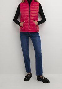 Czerwona pikowana kamizelka z zamkiem błyskawicznym i bocznymi kieszeniami, noszona na czarnej koszuli z długim rękawem, w połączeniu z niebieskimi dżinsami i czarnymi mokasynami.