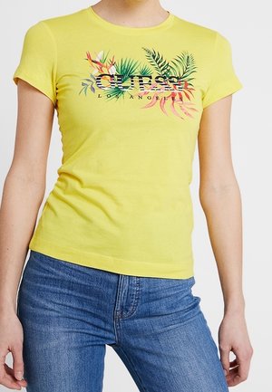 T-shirt jaune ajusté avec des feuilles tropicales colorées et le texte "GUESS LOS ANGELES", porté avec un jean bleu par une personne debout.
