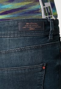 Jeans de mezclilla oscura con una cintura texturizada, que presentan una etiqueta que dice "Joven y Auténtico", y herrajes de acento rojo en el bolsillo.