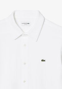 Weißes Baumwollpolohemd im slim fit Design mit klassischem Kragen und kleinem grünen Lacoste-Krokodil-Logo auf der linken Brust.