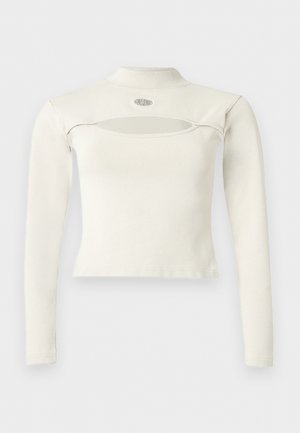 Top alb cu mâneci lungi, scurtat, cu guler înalt și o tăietură orizontală sub guler, cu un logo mic circular deasupra.