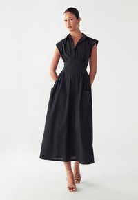 Črna midi obleka s kratkimi rokavi, ovratnikom na gumb, prilegajočim se pasom in stranskimi žepi; izdelana iz lahkega materiala z gladko teksturo.