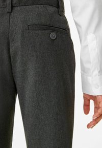 Pantalons ajustés gris foncé en tissu texturé, avec une poche arrière à bouton. Une manche de chemise blanche est visible à droite.
